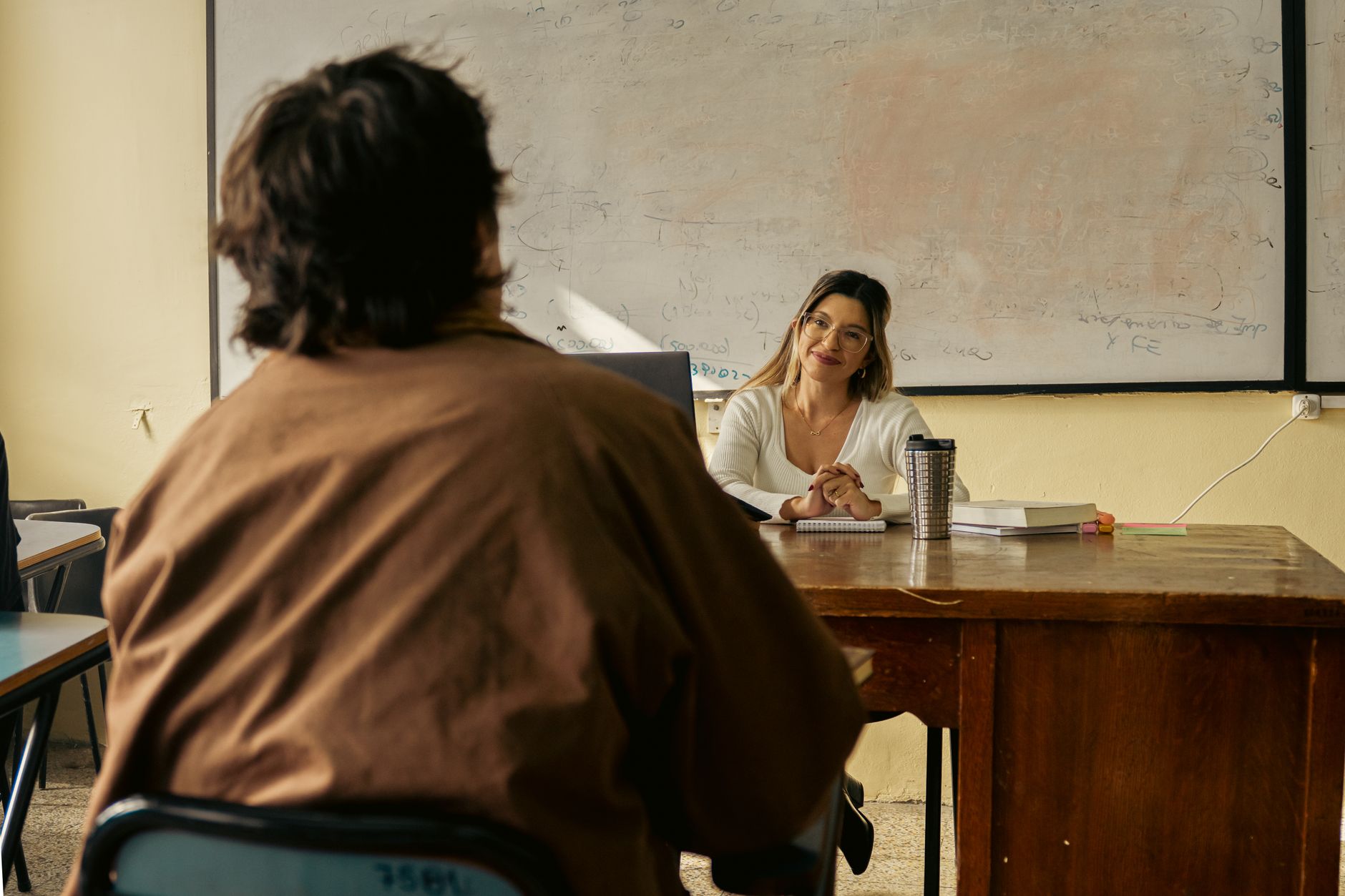 Professora sorridente conversa com aluno em sala de aula, quadro branco ao fundo e garrafa térmica na mesa.